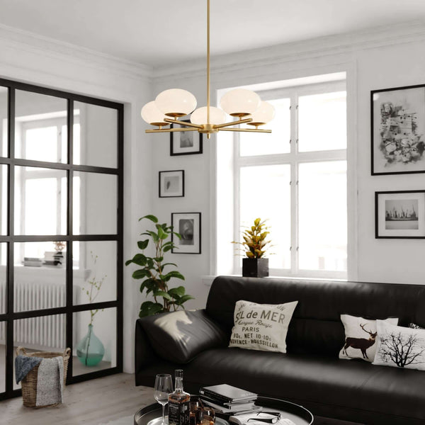 Sloane 27 inch 5 Light Chandelier in polished brass hanging above a modern living room with black sofa, wall art, and large windows, featuring smooth opal white glass globes creating warm diffused illumination.